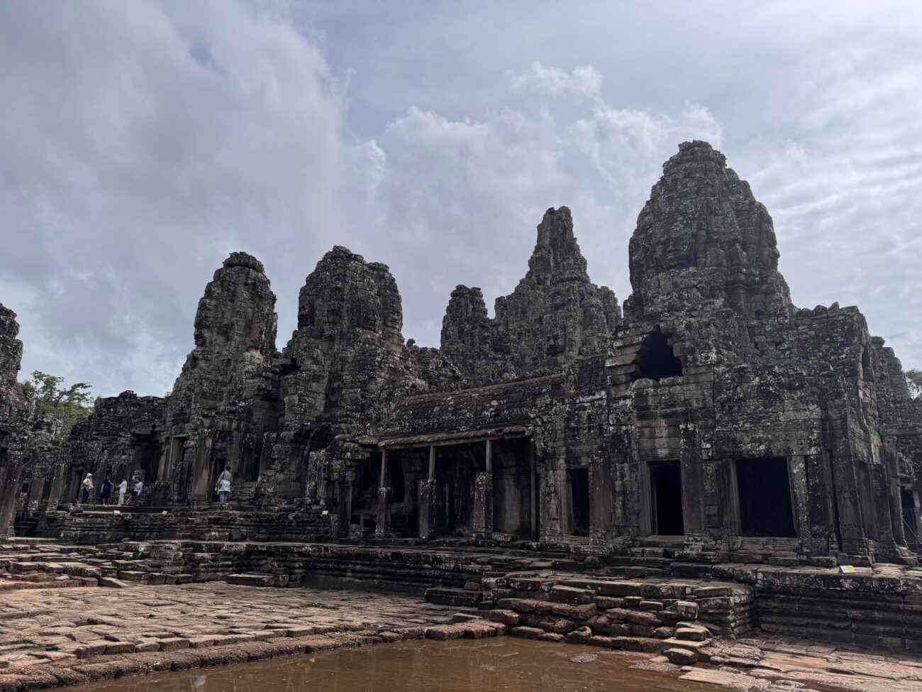 Eine Nahaufnahme der massiven geschnitzten Steintürme des Bayon Tempels im Angkor Thom Komplex in Kambodscha bedeckt mit Stein Gesichtern. Der verwitterte Steinboden und eine Wasserpfütze im Vordergrund spiegeln das Gebäude wider. Am unteren Rand stehen kleine Touristen für einen Größenvergleich.