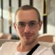 Ein Porträt von Marcel dem Autor des Blogs Alltagsbilanz. Er trägt eine Brille mit dunklem Gestell sowie ein weißes T Shirt und blickt freundlich direkt in die Kamera. Der Hintergrund ist leicht unscharf gehalten was den Fokus ganz auf seine Person lenkt.